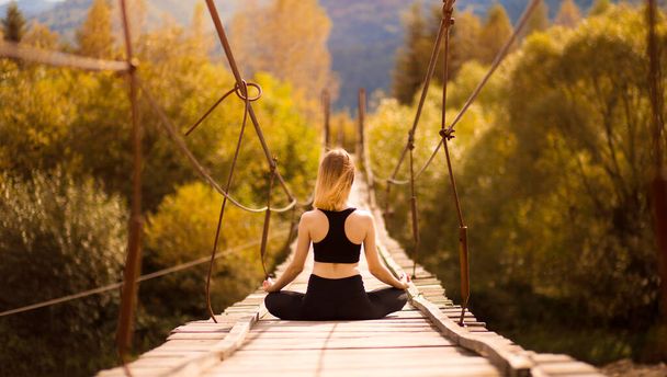 Vista posteriore della ragazza calma che pratica meditazione yoga seduta in posa di loto sul ponte sul fiume di montagna. Concetto di benessere.  - Foto, immagini