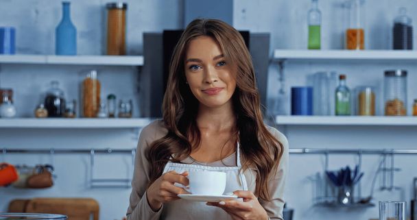 cheerful young woman in apron holding cup with coffee and saucer  - 写真・画像