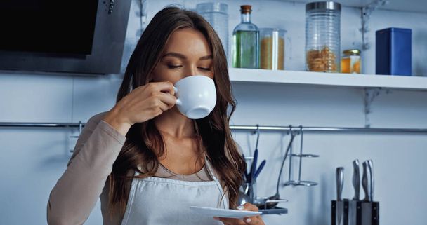 brunette young woman in apron drinking coffee and holding saucer  - Foto, imagen
