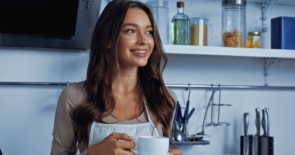 fröhliche junge Frau in Schürze mit Kaffeebecher und Untertasse  - Foto, Bild