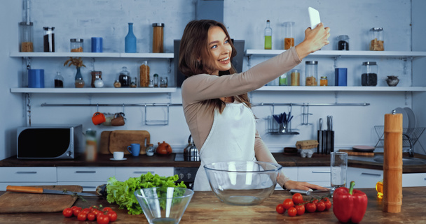 vrolijke vrouw in schort het nemen van selfie in de buurt ingrediënten op tafel - Foto, afbeelding