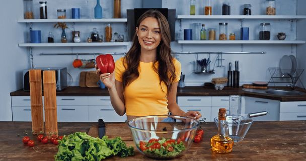 happy woman holding fresh bell pepper near ingredients on table - Photo, Image