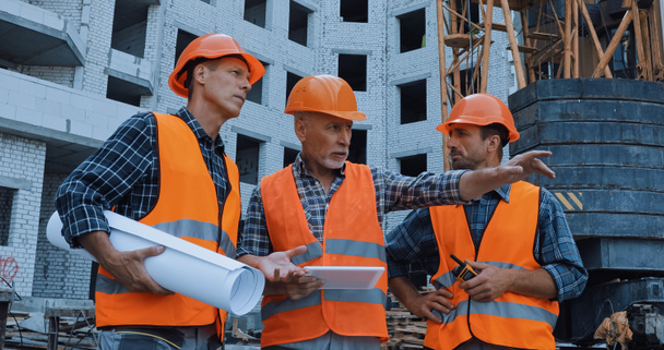 ώριμος εργαζόμενος που δείχνει με το χέρι και μιλάει κοντά σε συναδέλφους και γερανό κτίριο στο εργοτάξιο - Φωτογραφία, εικόνα