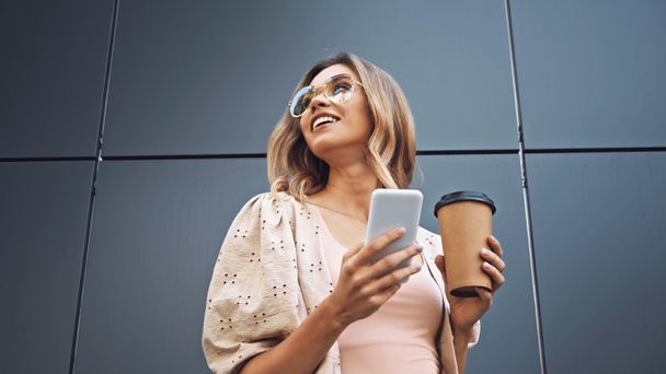 low angle view of blonde woman holding coffee to go and smartphone near building  - Photo, Image