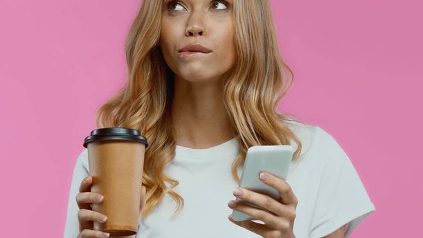 zamyślona kobieta za pomocą smartfona i trzymając kawę, aby przejść izolowane na różowy - Zdjęcie, obraz