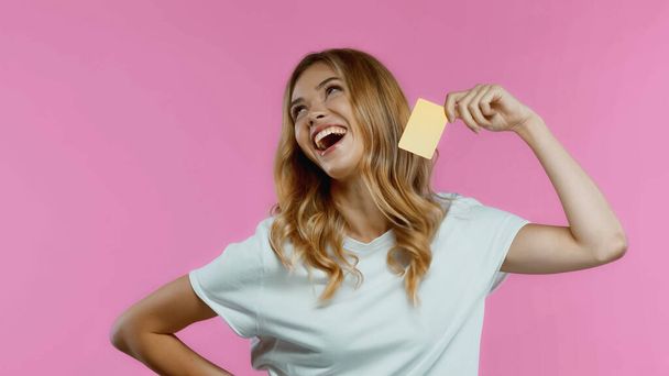 glückliche blonde Frau, die mit der Hand auf der Hüfte steht und Kreditkarte isoliert auf rosa hält  - Foto, Bild