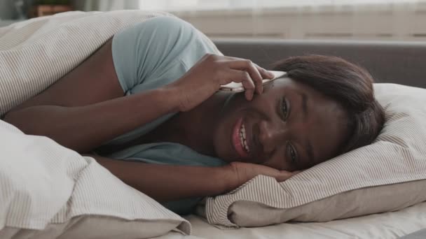 Encierro de una joven africana con pijamas azules acostada en la cama y hablando con una amiga por teléfono - Imágenes, Vídeo