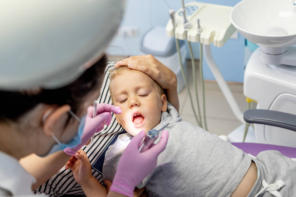 Mutter hält ein kleines Kind in einem Stuhl in einer hellen Zahnarztpraxis, Zahnarzt behandelt Zähne - Foto, Bild