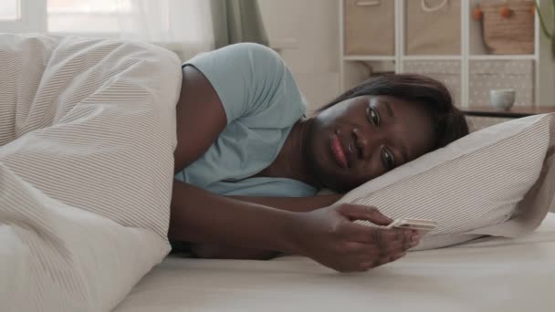 Bloqueo de una joven africana con pijamas azules acostada en la cama y mirando a través de fotos en red usando su teléfono - Metraje, vídeo