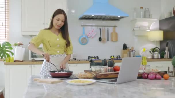 Egy gyönyörű ázsiai lány úgy készíti a reggelit, hogy megnézi, hogyan kell elkészíteni a laptopjával, boldog arccal. A saját házában. - Felvétel, videó