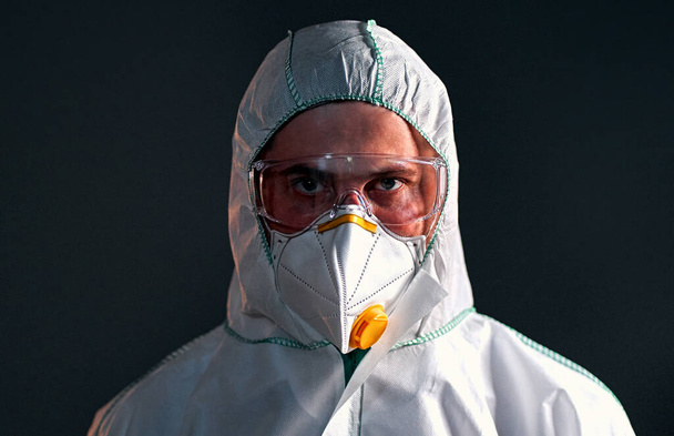 Male doctor or laboratory researcher is wearing a protective suit, a respirator and glasses are isolated on a dark gray background. - Photo, Image