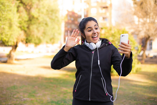 Молодая брюнетка спортивная девушка в наушниках прогуливаясь по парку с помощью смартфона и говоря привет очень счастлива - Фото, изображение