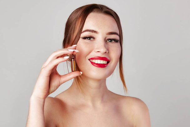 Frau hält Ampulle mit Serum für Haar- oder Hautpflege. Foto der attraktiven Frau mit Nasenohrring, roten Lippen und perfektem Make-up auf grauem Hintergrund. Schönheitskonzept - Foto, Bild