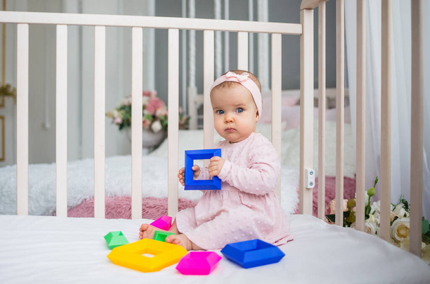 kaunis vauva tyttö vaaleanpunainen mekko ja otsanauha istuu vauvansängyssä ja leikkii opettavaisella lelulla - Valokuva, kuva