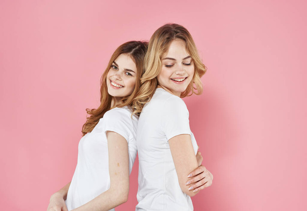 Two girlfriends in white T-shirts stand with their backs to each other on a pink background - Photo, Image
