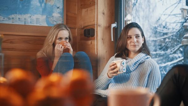 Καλύτεροι φίλοι περνούν τις χειμερινές διακοπές τους στη ζεστή ξύλινη καλύβα στο βουνό. Πίνοντας ζεστό ρόφημα κοντά στο μεγάλο παράθυρο - Φωτογραφία, εικόνα
