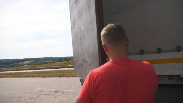Mężczyzna kierowca zamyka tylne drzwi ciężarówki na wsi. Ciężarówka zaparkowana przy drodze w słoneczny dzień. Piękny krajobraz w tle. Logistyka i koncepcja transportu. Zamknij Powolny ruch - Materiał filmowy, wideo