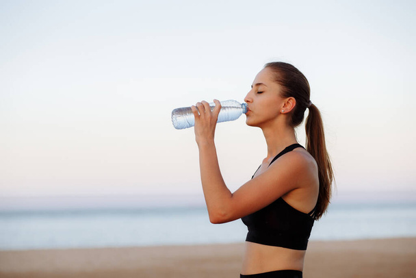 bella ragazza di sport in una cima e scarpe da ginnastica su una corsa del mattino beve l'acqua da una bottiglia - Foto, immagini