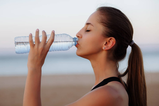 hermosa chica deportiva en una parte superior y zapatillas de deporte en una carrera de la mañana bebe agua de una botella - Foto, Imagen