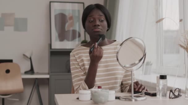 Μέτριο πλάνο της νεαρής ελκυστική Αφροαμερικανή γυναίκα κάθεται στο τραπέζι καθρέφτη στο σπίτι και την εφαρμογή σκόνη προσώπου χρησιμοποιώντας βούρτσα πριν βγείτε έξω - Πλάνα, βίντεο