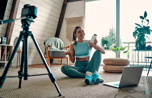 Atletische vrouwelijke blogger in sportkleding zittend op de vloer met halters en een laptop en met een potje sportvoeding proteïnen aan de camera thuis in de woonkamer. Sport en recreatie. - Foto, afbeelding