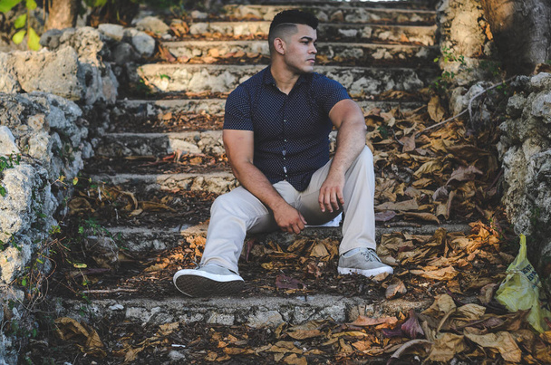 White-skinned male model of Venezuelan origin in the Dominican Republic. sexy man in jeans in the park outdoors with orange walks with natural sunlight among the leaves sitting on the stairs - Photo, Image