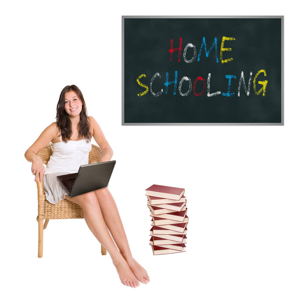 Smiling schoolgirl with a huge pile of books and a laptop in front of a blackboard, homeschooling is written on the board, isolated on white studio background - Photo, Image