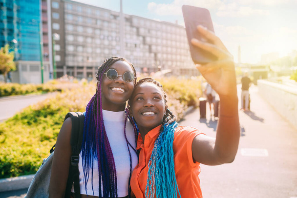 Duas irmãs negras ao ar livre tirando selfie usando a luz de fundo do smartphone sorrindo e rindo com boas vibrações positivas - Foto, Imagem