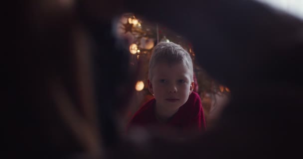 Frame portrét roztomilé blonďaté dítě pózovat na focení na Silvestra zpomalit. Ruční video záběr fotografa bez tváře střelba šťastný chlapec sedí u vánočního stromečku uvnitř.  - Záběry, video