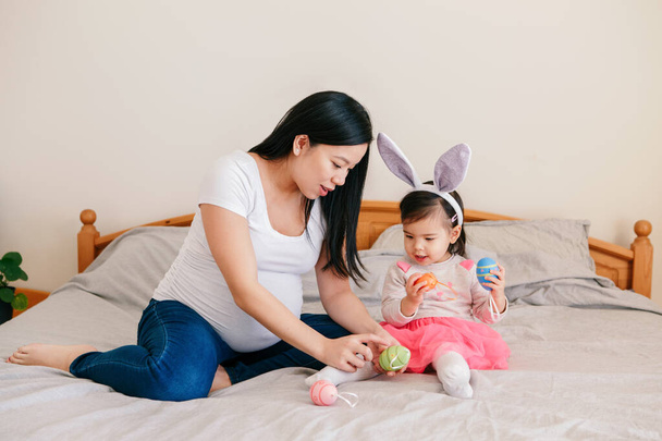 Feliz Páscoa. Mãe grávida chinesa asiática com bebê menina brincando com ovos de Páscoa coloridos na cama em casa. Criança e pai celebrando as tradicionais férias cristãs. - Foto, Imagem
