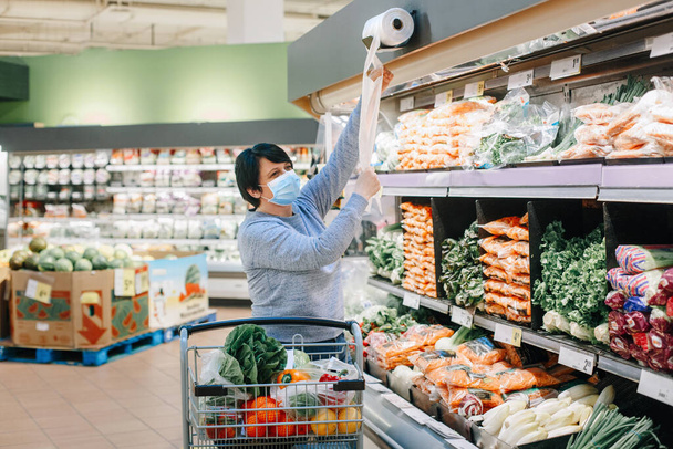 Obchod s potravinami. Žena středního věku s obličejovou maskou strhává plastovou průhlednou jednorázovou tašku v obchodě s potravinami na nákup potravinové zeleniny. Nový normální na coronavirus covid-19. - Fotografie, Obrázek