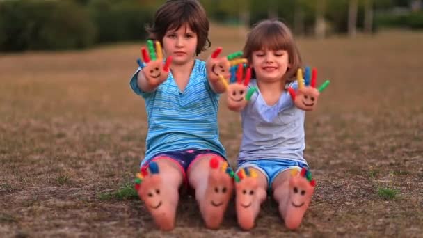 Két vicces kislány ül a füvön és integet festett kezekkel és lábakkal vicces arcokba.  - Felvétel, videó