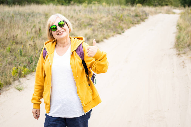 Ευτυχισμένη ηλικιωμένη γυναίκα με γυαλιά ηλίου και κίτρινη κουκούλα, που δείχνει τον αντίχειρά της σαν χειρονομία, συνιστώ. Backpacker χαμογελώντας κοιτάζοντας κάμερα, στέκεται στο μονοπάτι σε εξωτερικούς χώρους δασών.Απολαμβάνοντας ενεργό ταξίδια - Φωτογραφία, εικόνα