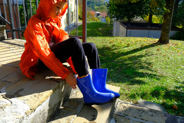 Lapsi oranssissa sadetakissa laittamassa sinisiä rubiinisaappaita takapihalle. Vaatteet sateinen sää - Valokuva, kuva