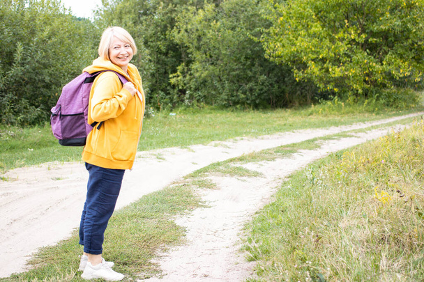 Gelukkige senior vrouw reiziger in gele hoodie met rugzak, lachend op pad in groen bos buiten. Actief leven van gepensioneerden. Geniet van weekend of vakantie. Kopieerruimte. - Foto, afbeelding