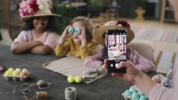Toma de fotos de una mujer irreconocible sosteniendo un teléfono móvil y tomando fotos de niños lindos en el taller de Pascua posando con sombreros de paja decorados con flores y huevos teñidos - Imágenes, Vídeo