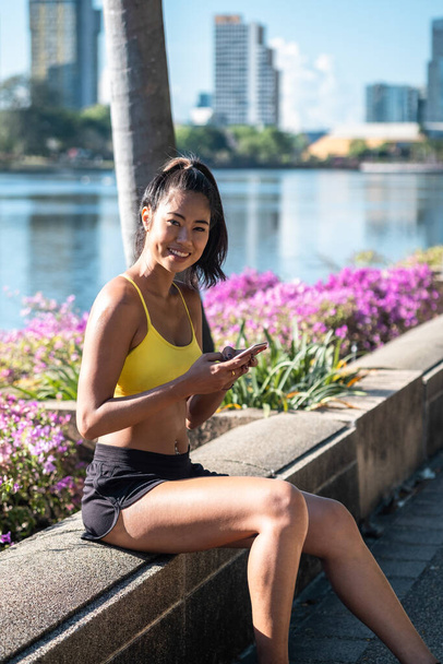 スポーツウェアの美しい笑顔の女性は、カメラを見て、携帯電話を保持し、訓練後に休息。ワークアウト後のパフォーマンスを追跡するための携帯電話フィットネスアプリを使用してスポーティアジアの女性 - 写真・画像