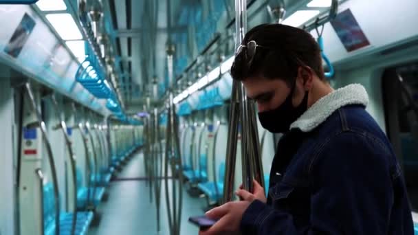 Un joven con máscara negra de pie en un tren subterráneo vacío y mirando su teléfono - Imágenes, Vídeo