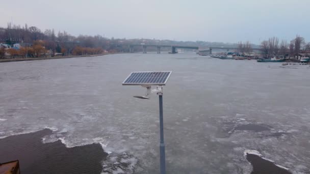 Klein zonnepaneel op stalen voetgangersbrug door rivier met smeltend ijs in koude tijd - Video