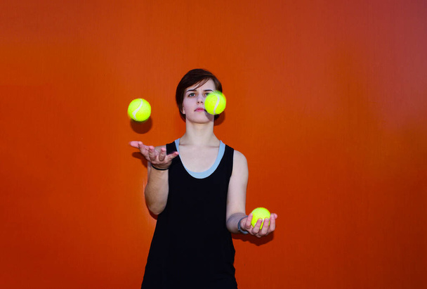 Молодая девушка жонглирует теннисными мячами на оранжевом фоне. Спорт и отдых - Фото, изображение