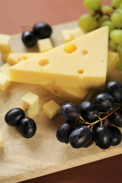 マスター・チーズだ。茶色の背景に木の板の上にチーズのマダムやマスダマー、緑と黒のブドウのピース。乳製品健康的でおいしいスナックスイスのマサダムチーズ - 写真・画像