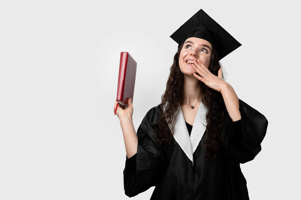 Student with book in graduation robe and cap ready to finish college. Future leader of science. Academician young woman in black gown smiling. - Photo, Image