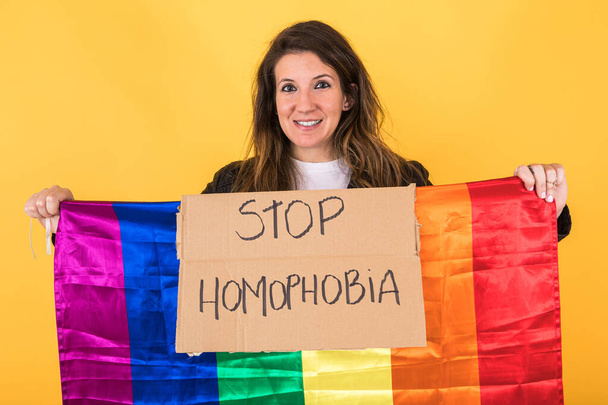 Серйозна гомосексуальна жінка тримає знак з закликом "Стоп-гомофобія" і гей-райдужний прапор на жовтому тлі. - Фото, зображення