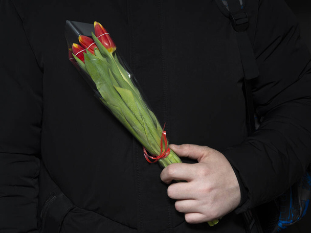 Ein Mann hält einen kleinen Strauß Tulpen in Geschenkpapier mit einer roten Schleife in der Hand vor der Brust. Das Konzept der Glückwünsche zum 8. März, Frühlingsfest, Muttertag oder Geburtstag. - Foto, Bild