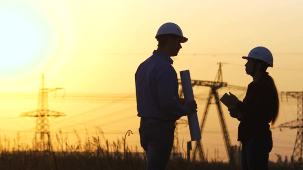Power engineers are discussing the work plan. Two engineer standing on field with electricity towers at sunset. Silhouette of engineers looks at construction of high-voltage power. Engineering team - Footage, Video