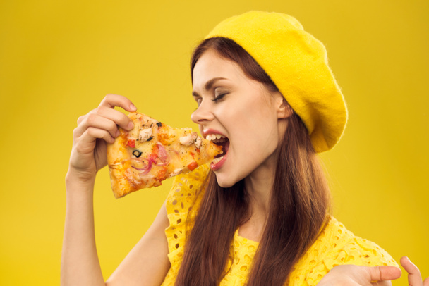 Woman in yellow hat with a piece of pizza in her hands on a yellow background cropped view of emotion model - Photo, Image