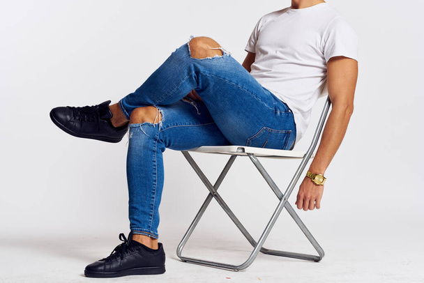 un hombre en jeans y una camiseta se sienta en una silla en una vista lateral de fondo claro - Foto, imagen
