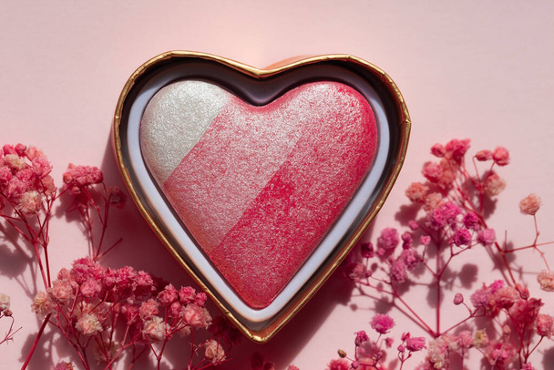 Коробка компактного порошка в форме сердца на розовом бумажном фоне с цветами гипсофилы. Вид сверху. Косметический продукт для щек. Запеченный румянец крупным планом - Фото, изображение