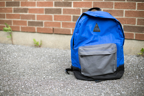 Iskolai hátizsák az úton. Kék fiú hátizsákja a járdán az iskola vörös téglafalánál a háttérben. Vissza az iskolai oktatási koncepcióhoz. - Fotó, kép
