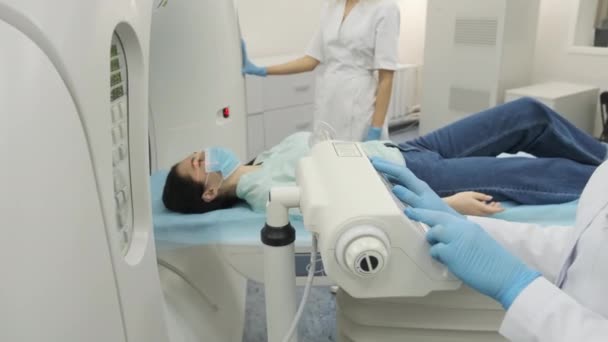 Une patiente subit une tomodensitométrie ou une IRM sous la supervision de deux radiologues qualifiés dans une clinique médicale moderne. Patient allongé sur une table de tomodensitométrie ou d'IRM, se déplaçant à l'extérieur de la machine - Séquence, vidéo